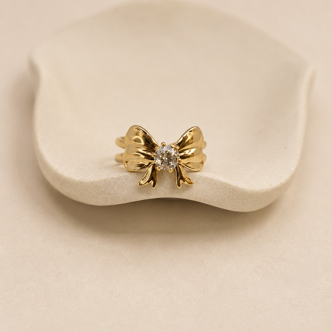 A gold bow-shaped ring with a clear central gemstone, displayed on a small white ceramic stand against a warm beige background.