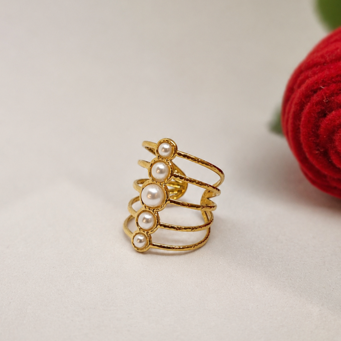 A gold-plated multi-band ring decorated with five white faux pearls, displayed on a light beige surface next to a red rose.