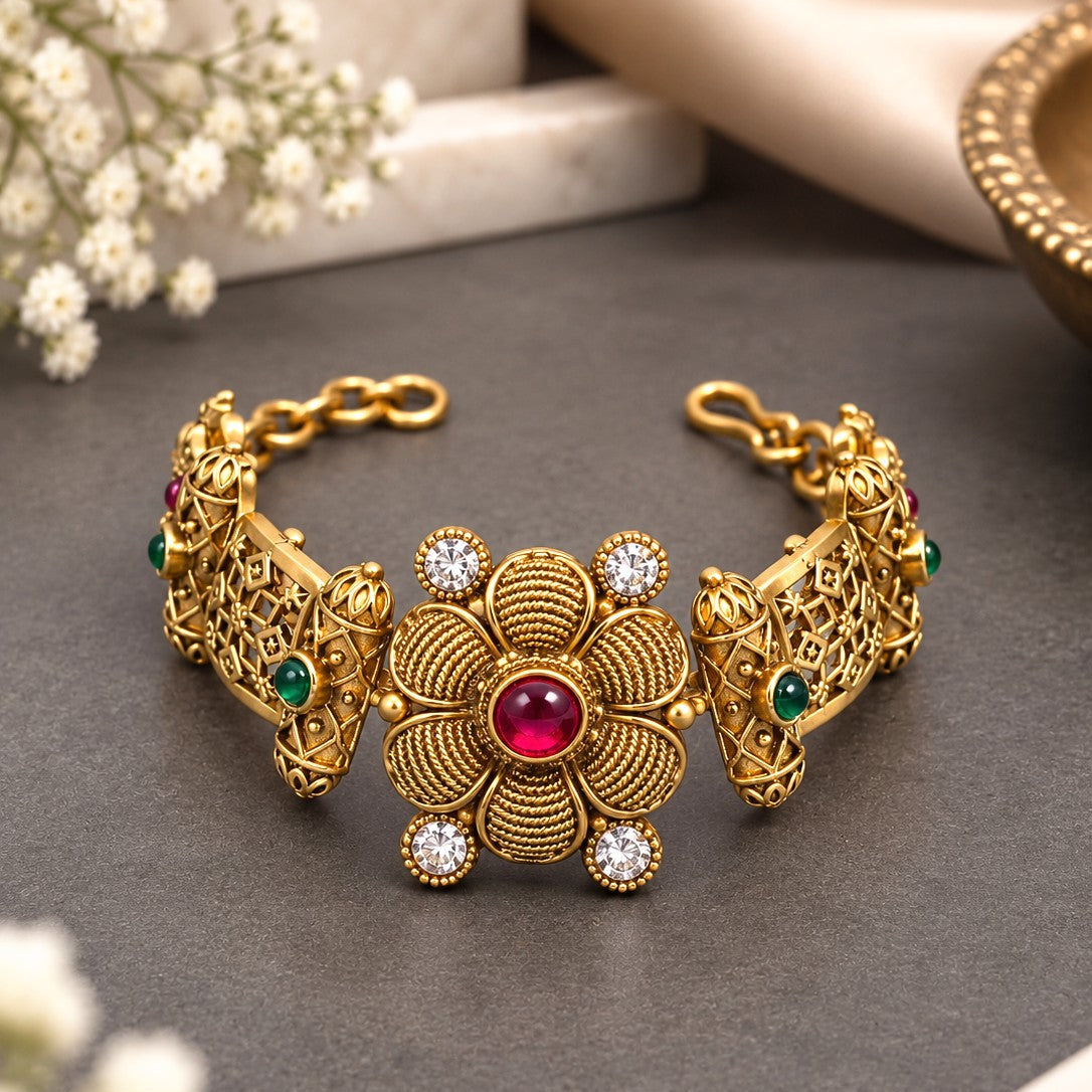 An ornate gold-toned traditional cuff bracelet with a central floral motif, a red center stone, and small green and clear accent stones, displayed on a dark gray surface with decorative flowers and fabric in the background.