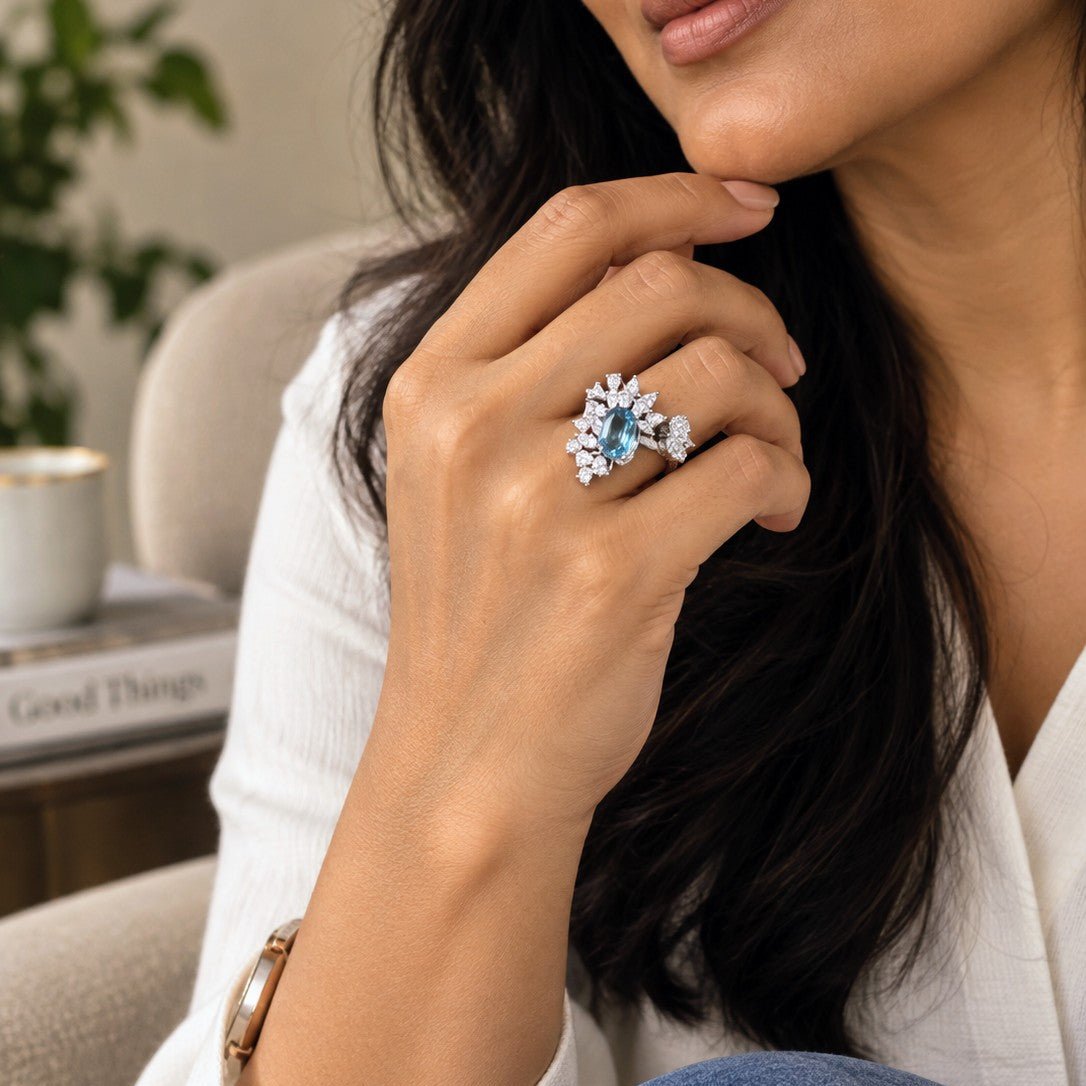 A close-up lifestyle shot of a woman wearing a silver-toned ring with a large light blue round center stone and smaller clear accent stones. She is wearing a white textured top and blue jeans, with a neutral indoor background featuring a chair and a plant.