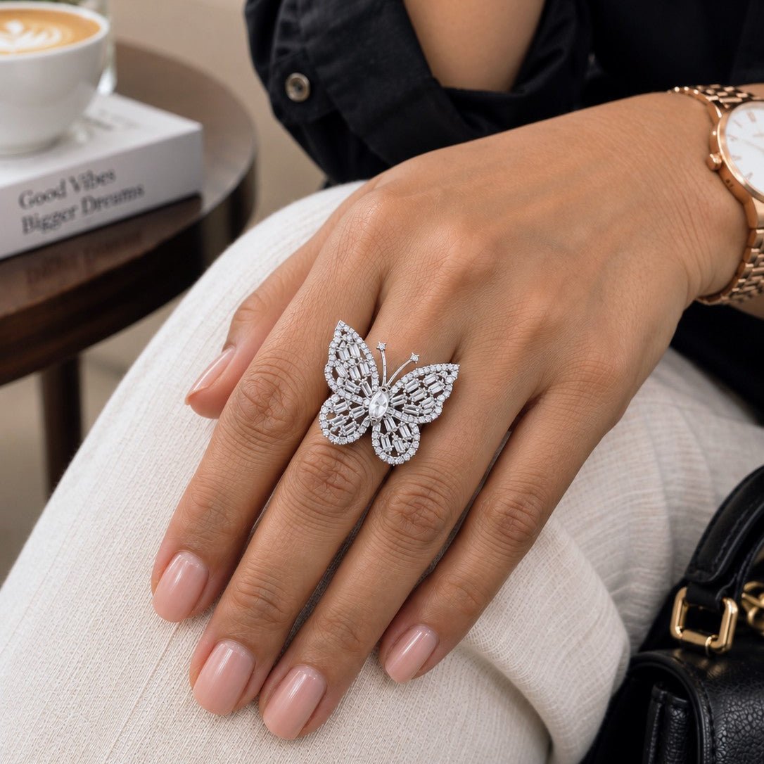 A close-up lifestyle shot of a hand wearing a sparkling silver butterfly ring, styled with a black shirt, cream pants, and a gold watch, with a coffee cup and book in the background.