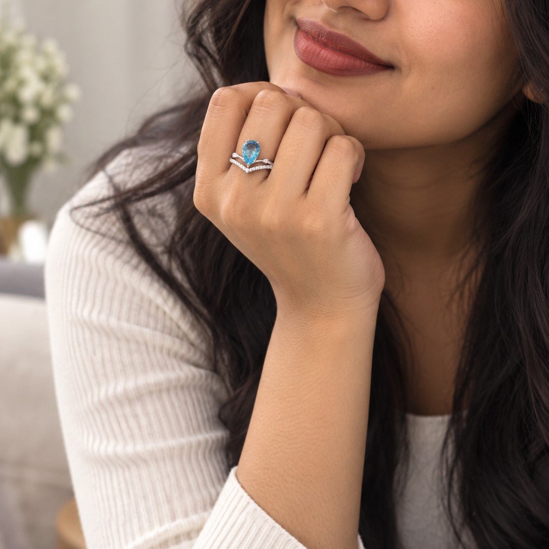 A close-up lifestyle shot of a woman wearing a silver ring with a blue teardrop-shaped gemstone and small clear accent stones, resting her chin on her hand.