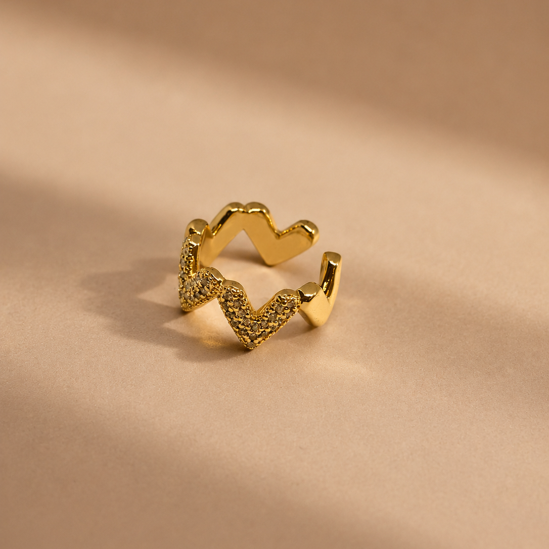 A gold-toned open ring band featuring a wavy, zigzag pattern with textured pavé detailing on one side and smooth polished edges on the other, resting on a warm beige surface with soft shadows.