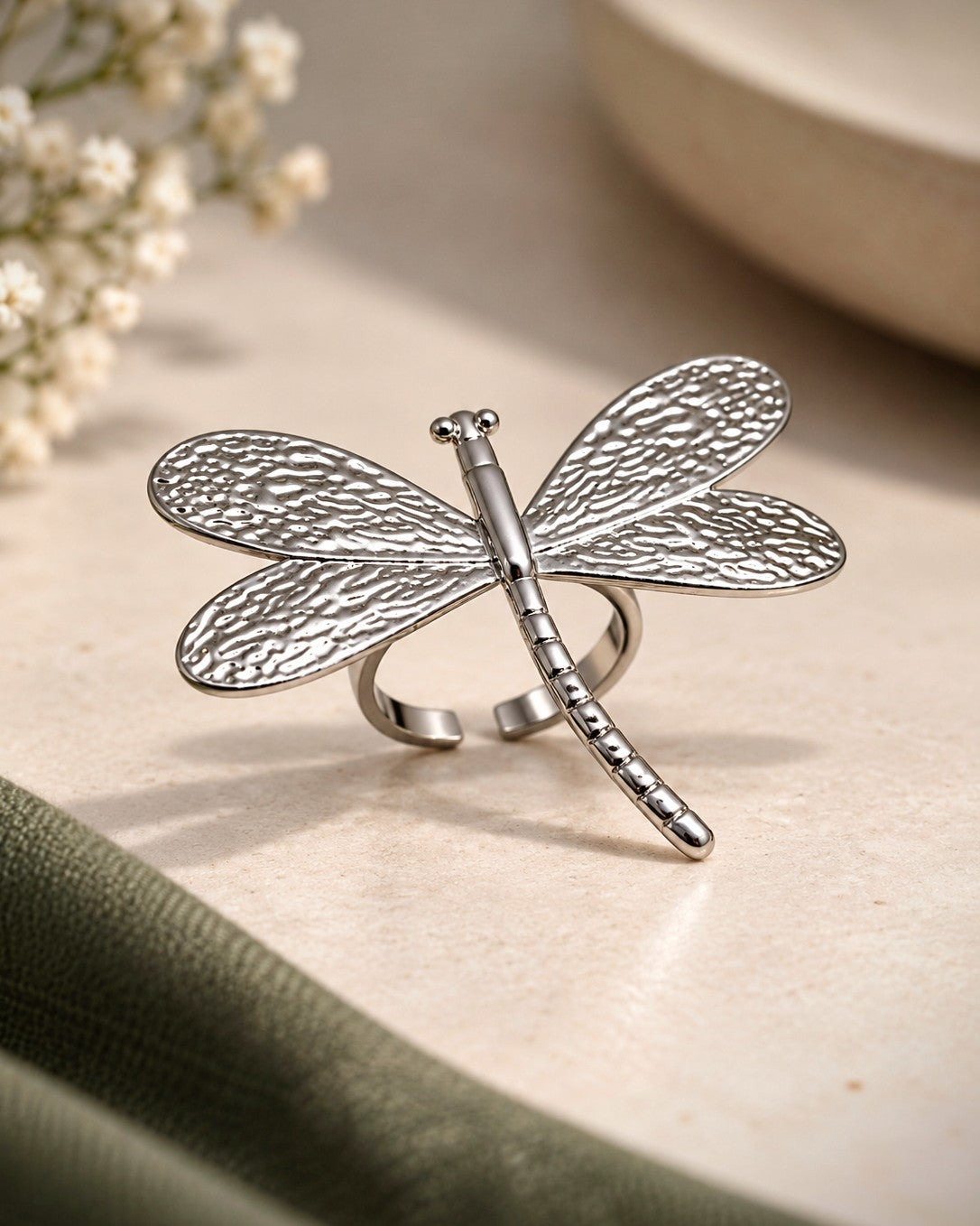A silver dragonfly ring with textured wings and a segmented body, displayed on a light beige surface with soft-focus floral and fabric props.