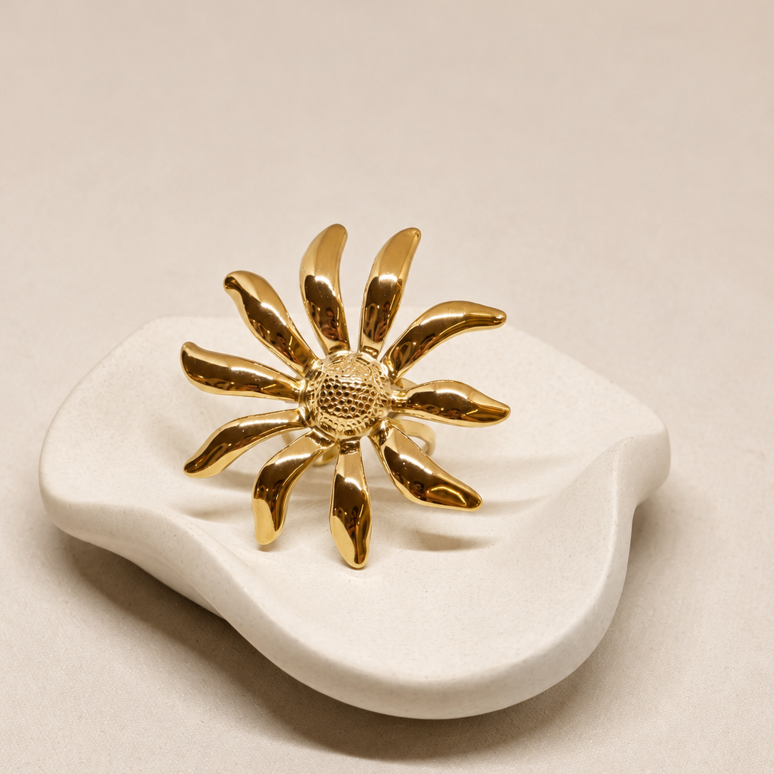 A gold flower-shaped ring displayed on a small white ceramic dish against a warm beige background.