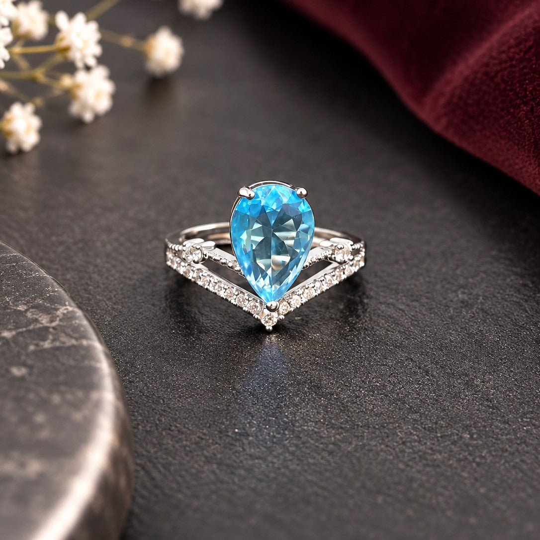 A silver ring featuring a pear-cut blue gemstone and a V-shaped pavé band, displayed on a dark textured surface with a burgundy fabric and small white flowers in the background.
