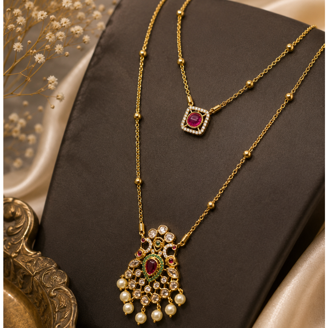 Two gold-toned traditional Indian necklaces displayed on a dark brown jewelry stand. The necklace at the top has a square pendant with a red stone and clear stones, while the larger necklace at the bottom features an ornate hand-shaped pendant with a red stone, green stone, clear stones, and dangling white faux pearls.