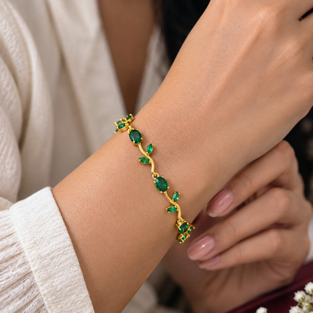 A gold bracelet with green leaf-shaped stones worn on a woman's wrist, shown in a close-up lifestyle shot with a cream-colored top and a small red jewelry box in the background.