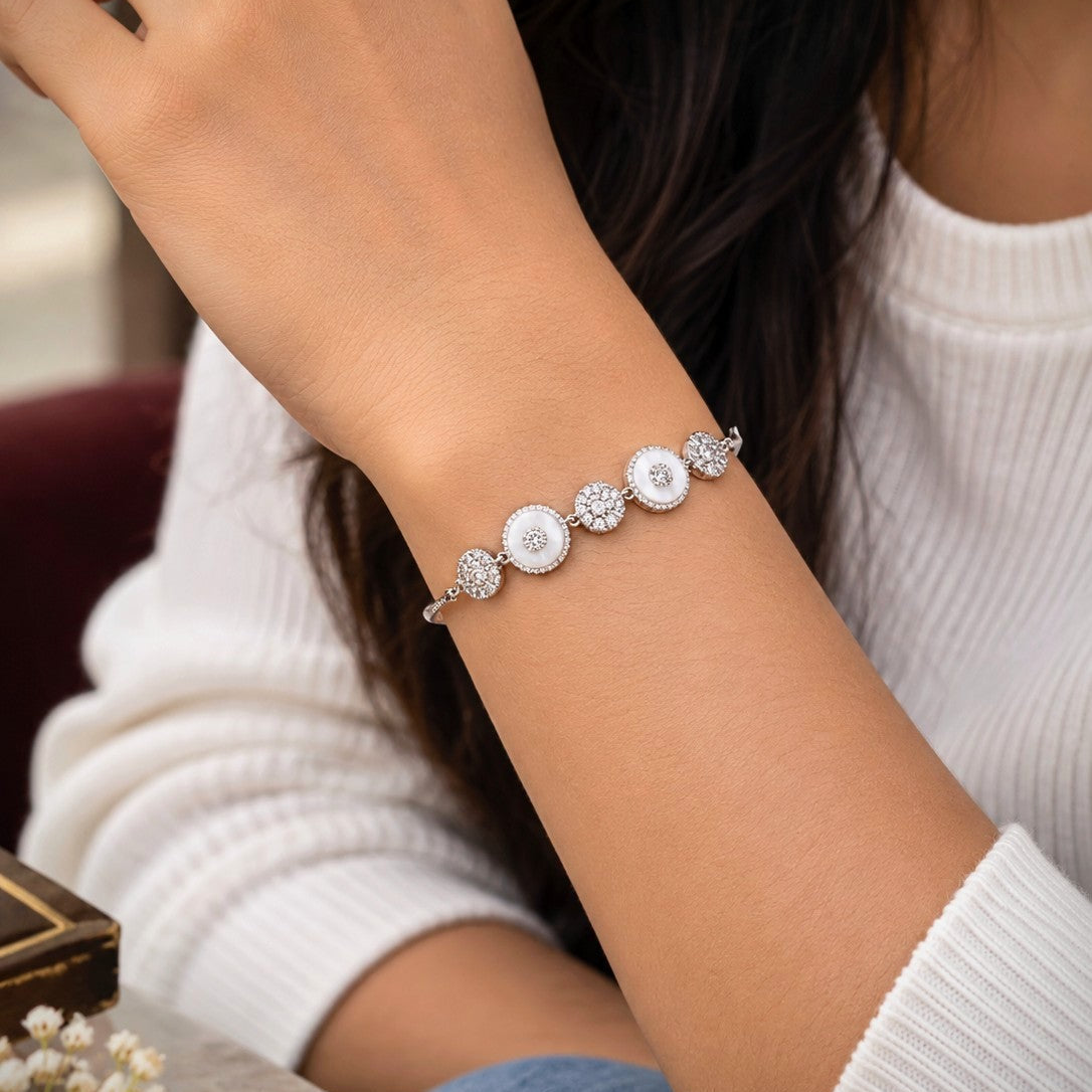 A close-up of a person's wrist wearing a delicate silver bracelet with three round white pearl-like beads and small clear crystal details, paired with a white ribbed sweater.