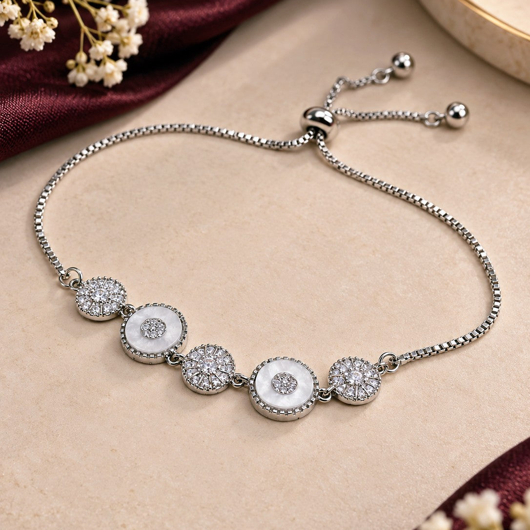 A silver chain bracelet with five circular white inlay and crystal-encrusted medallions, displayed on a beige surface with dark red fabric and small white flowers.