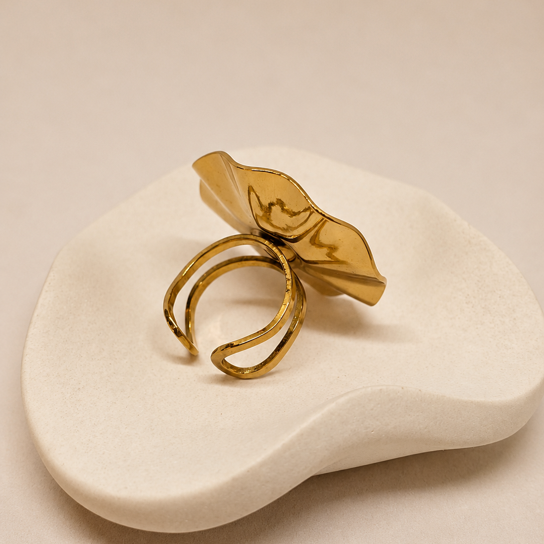 A gold-toned ring with a sculptural, organic leaf-like top and two curved bands, displayed on a small white ceramic dish against a warm beige background.