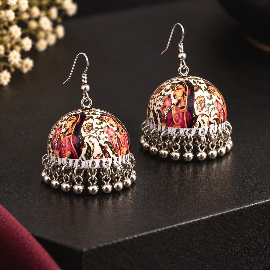 A pair of traditional Indian jhumka earrings with silver-toned filigree borders and domed charms featuring a colorful floral and paisley pattern in red, pink, gold, and black, displayed on a dark textured surface.