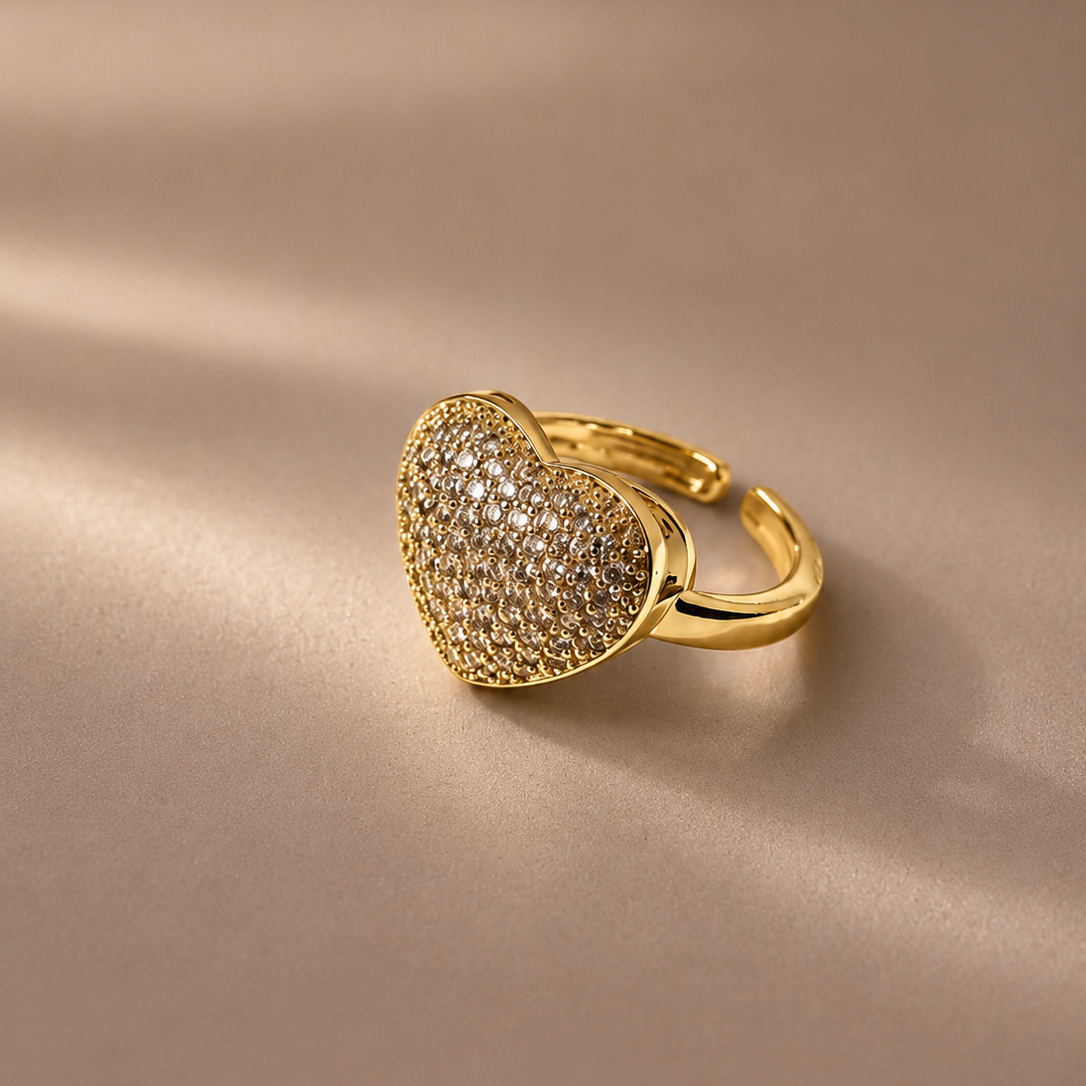 A gold heart-shaped ring with a pavé-set clear stone center, resting on a soft beige satin surface with elegant shadows.