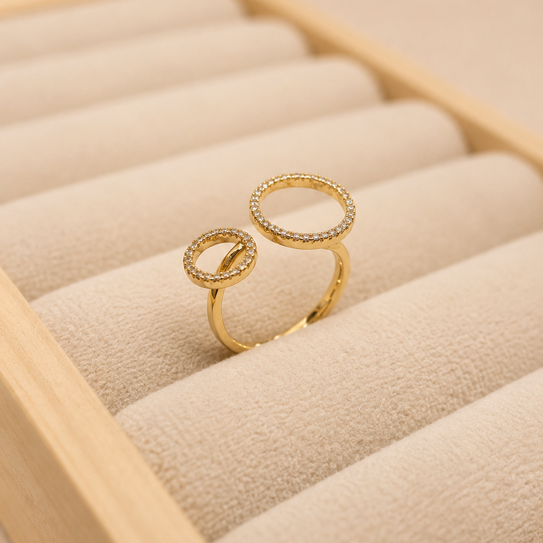 A pair of gold-toned open ring rings with pavé-set clear stones, displayed on a beige jewelry box cushion.