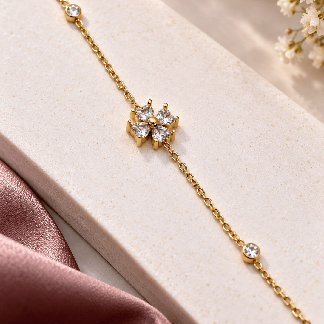 A delicate gold chain bracelet with a central flower-shaped cluster of clear gemstones and small accent stones, displayed on a light beige textured block with pink satin and white flowers in the background.