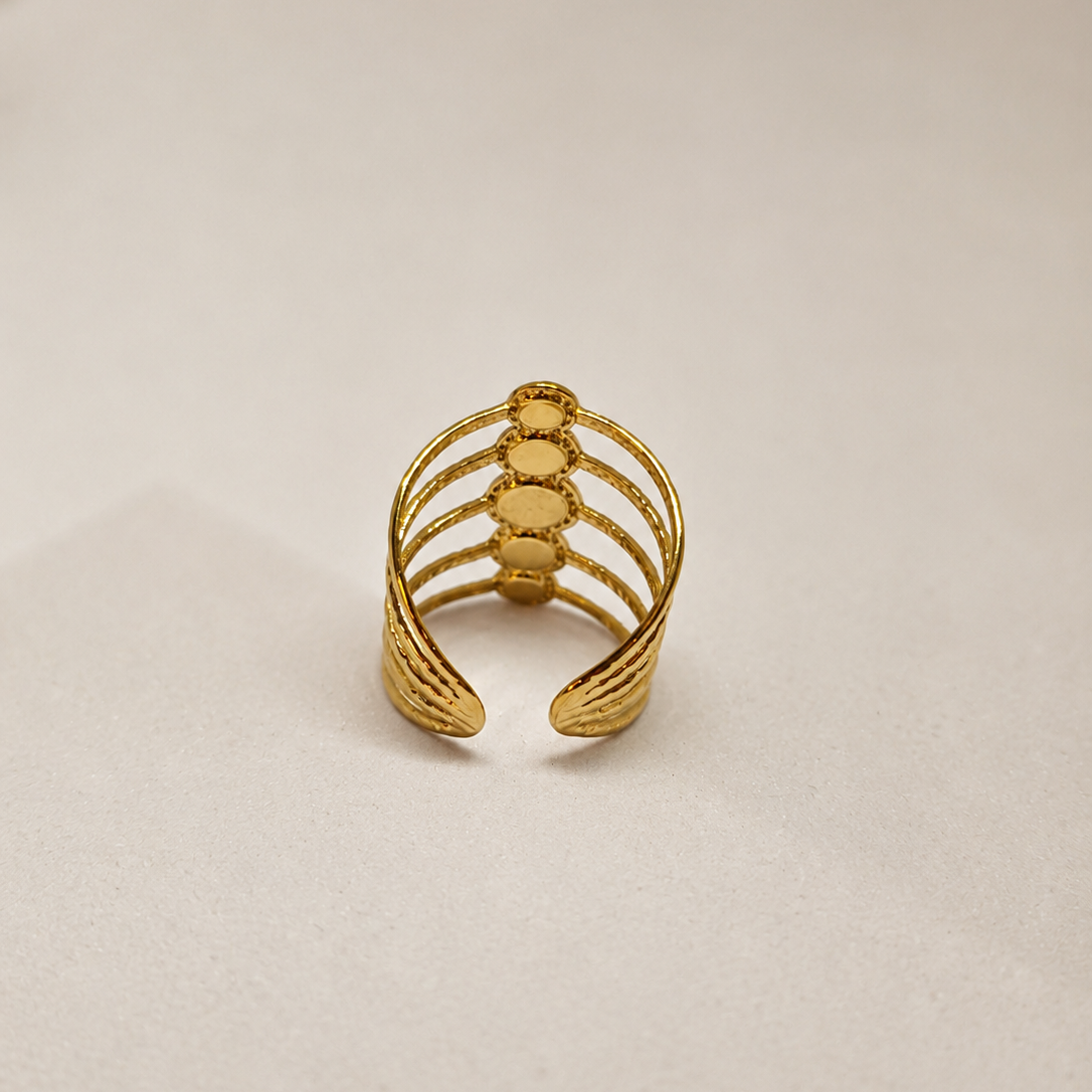 A gold-toned open ring with a ribbed textured design and a central vertical row of oval details, photographed on a plain light beige surface.