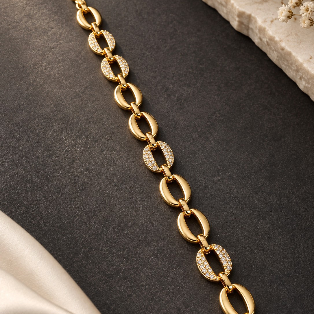 A gold chain bracelet with alternating polished oval links and oval links set with small clear stones, displayed diagonally on a dark textured surface with decorative fabric and stone props.