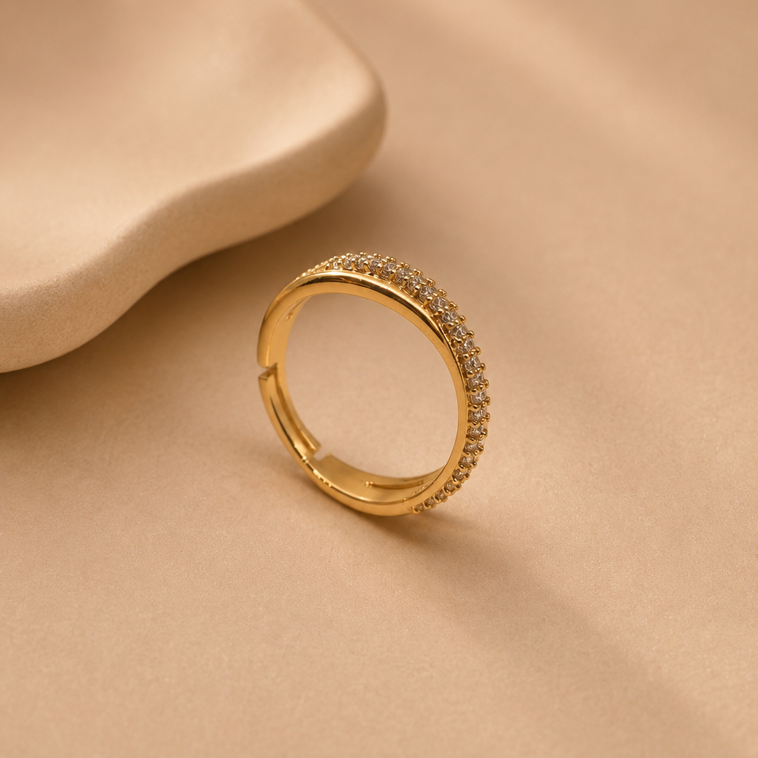 A gold ring with a textured, pavé-set stone band, resting on a warm beige textured surface next to a matching beige abstract prop.
