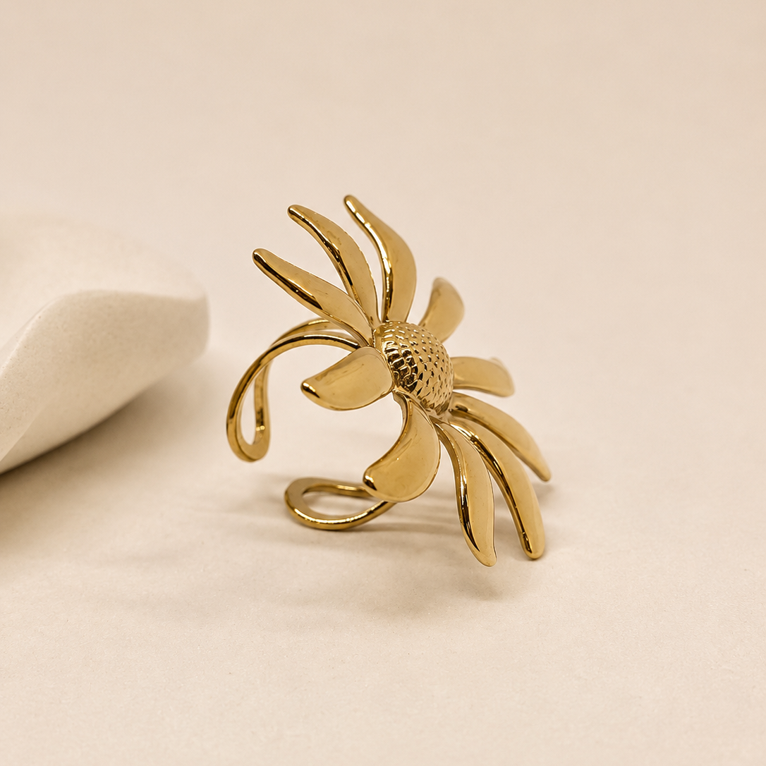 A gold flower-shaped ring with a textured center and curved petals, displayed on a warm beige surface next to a white decorative stone.