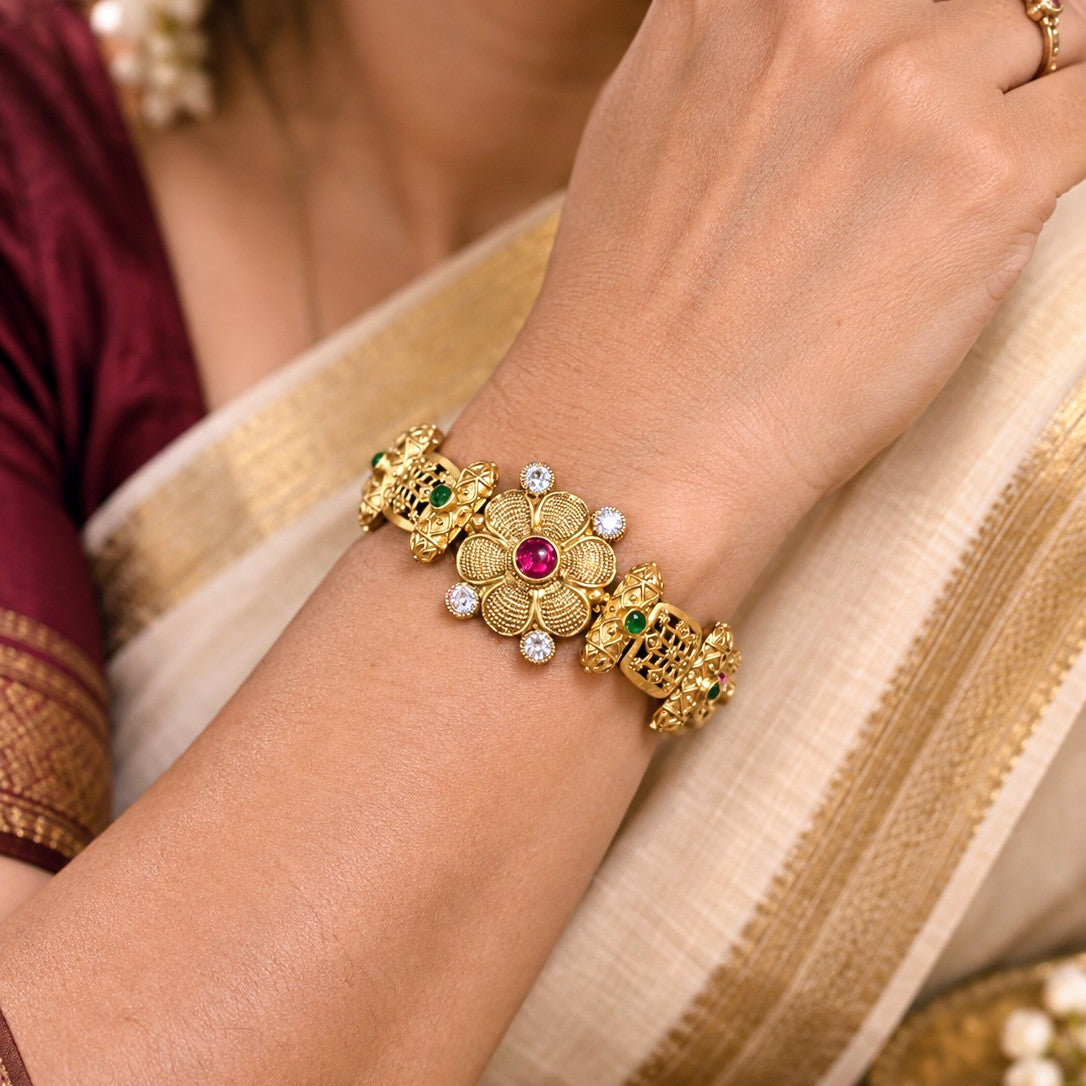 A close-up of a woman's wrist wearing an ornate traditional Indian gold bracelet with a central floral motif, a red ruby stone, and small white pearl-like accents. The background shows a cream and gold saree and part of a maroon blouse.