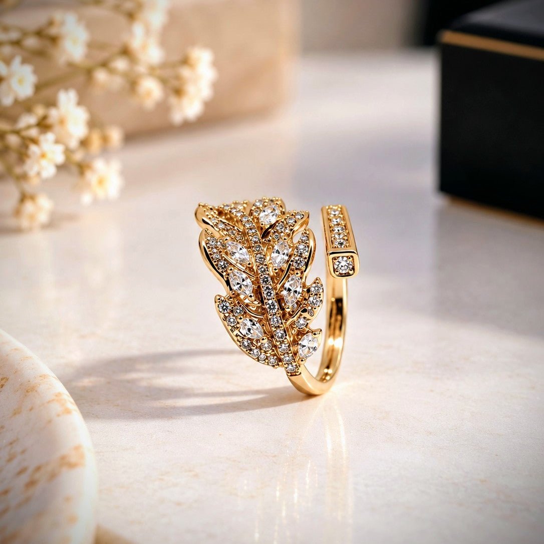 A gold ring featuring a leaf-shaped pavé setting with clear stones, displayed on a light marble surface with soft-focus decorative props.