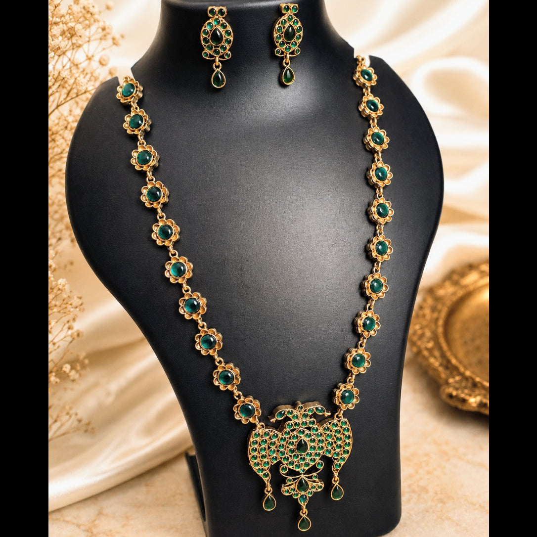 Gold and emerald green jewelry set displayed on a black mannequin bust, including a necklace with a large ornate pendant and matching drop earrings, set against a cream satin background with decorative dried flowers.