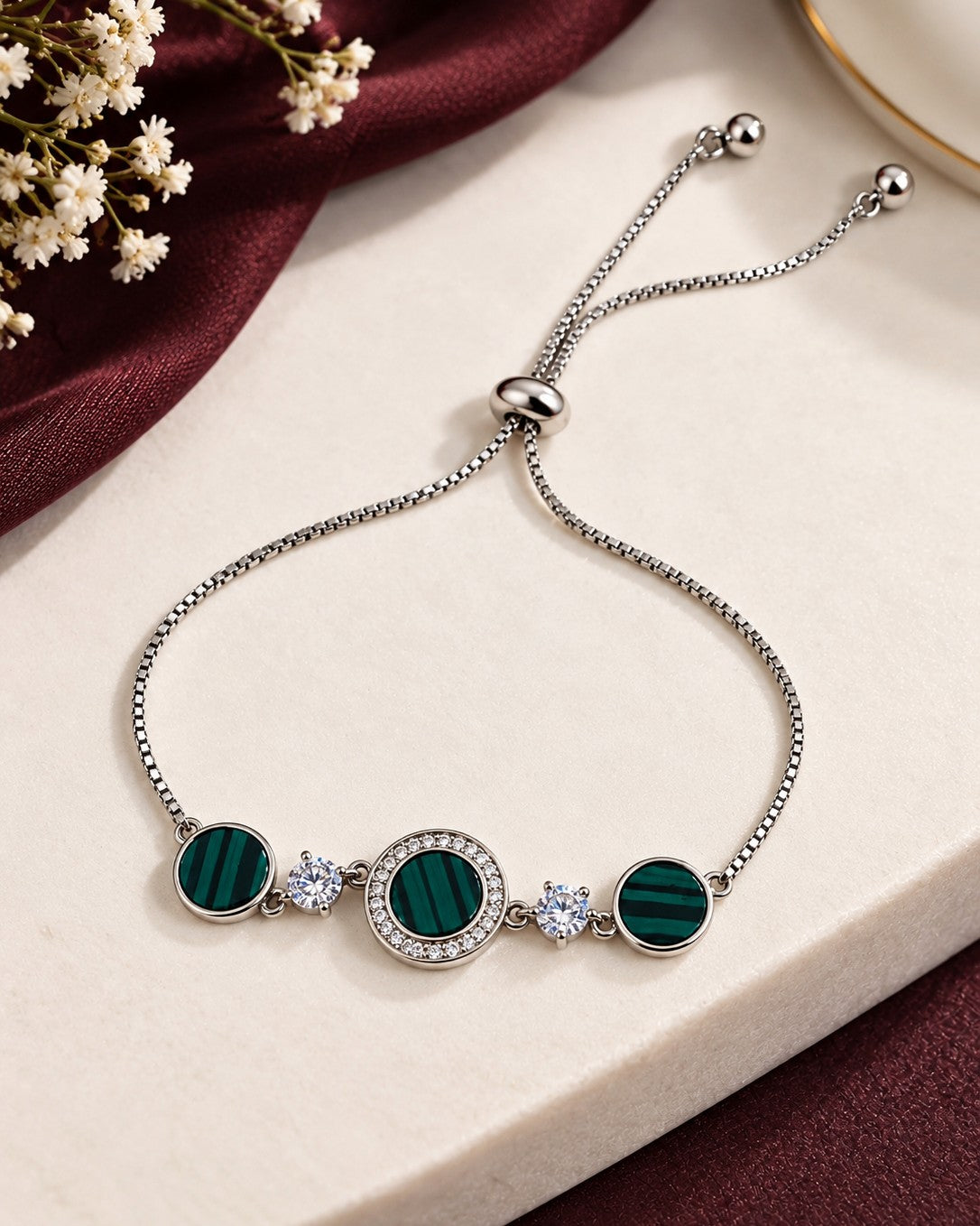 A silver adjustable chain bracelet with three oval green malachite stone settings and small clear crystal accents, displayed on a cream surface with burgundy fabric and dried flowers.