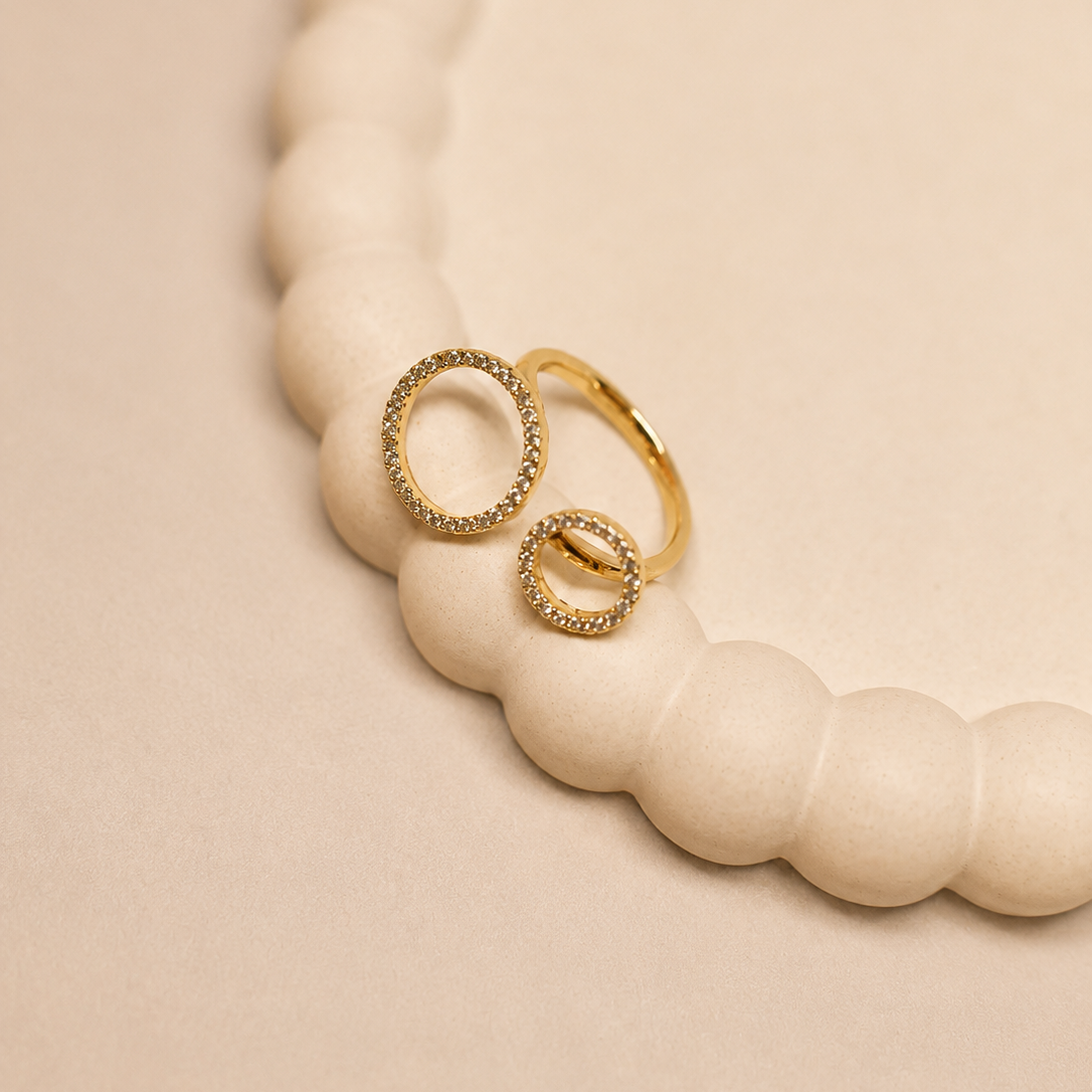 Two gold rings with textured and smooth oval designs, displayed on a cream-colored beaded tray against a warm beige background.