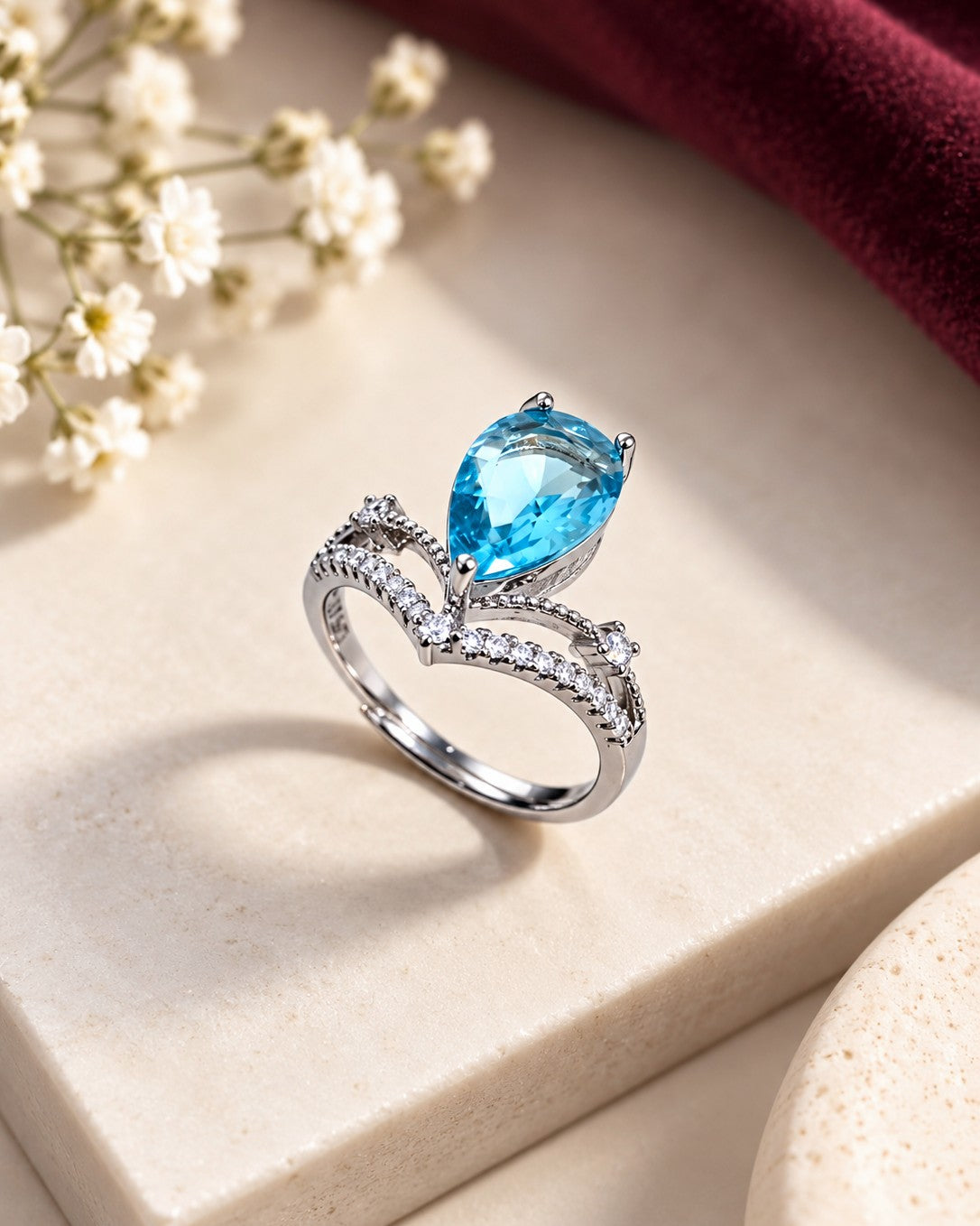 A silver ring featuring a large pear-cut blue gemstone and a curved, pavé-set silver band, styled on a beige stone block with white baby's breath flowers and a dark red fabric background.