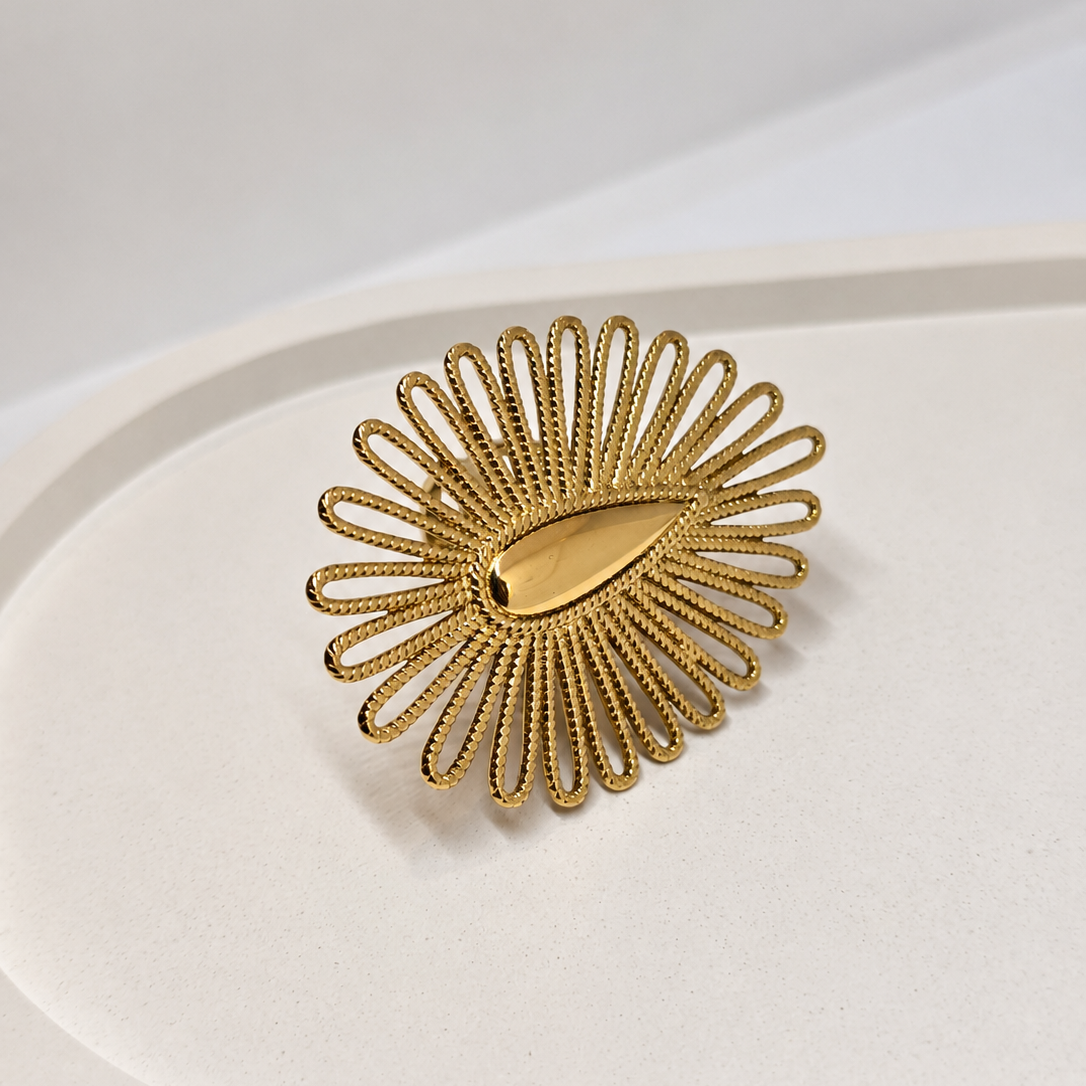 A gold-toned sunburst ring with a textured, openwork design and a smooth teardrop-shaped center, displayed on a minimalist white surface.