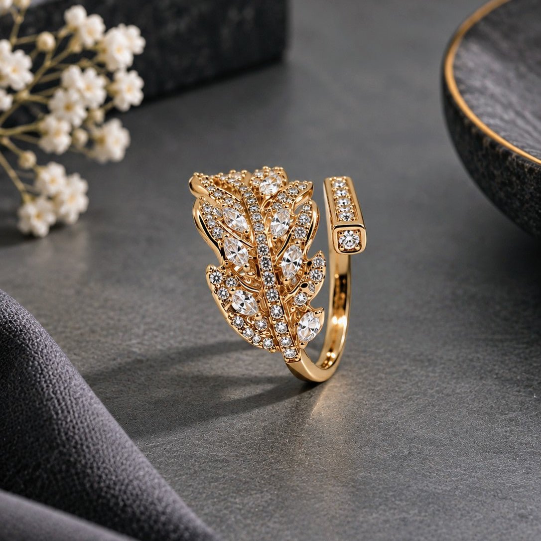 A gold leaf-shaped ring adorned with clear pavé gemstones, displayed on a dark textured surface with decorative dried flowers and a dark stone bowl in the background.
