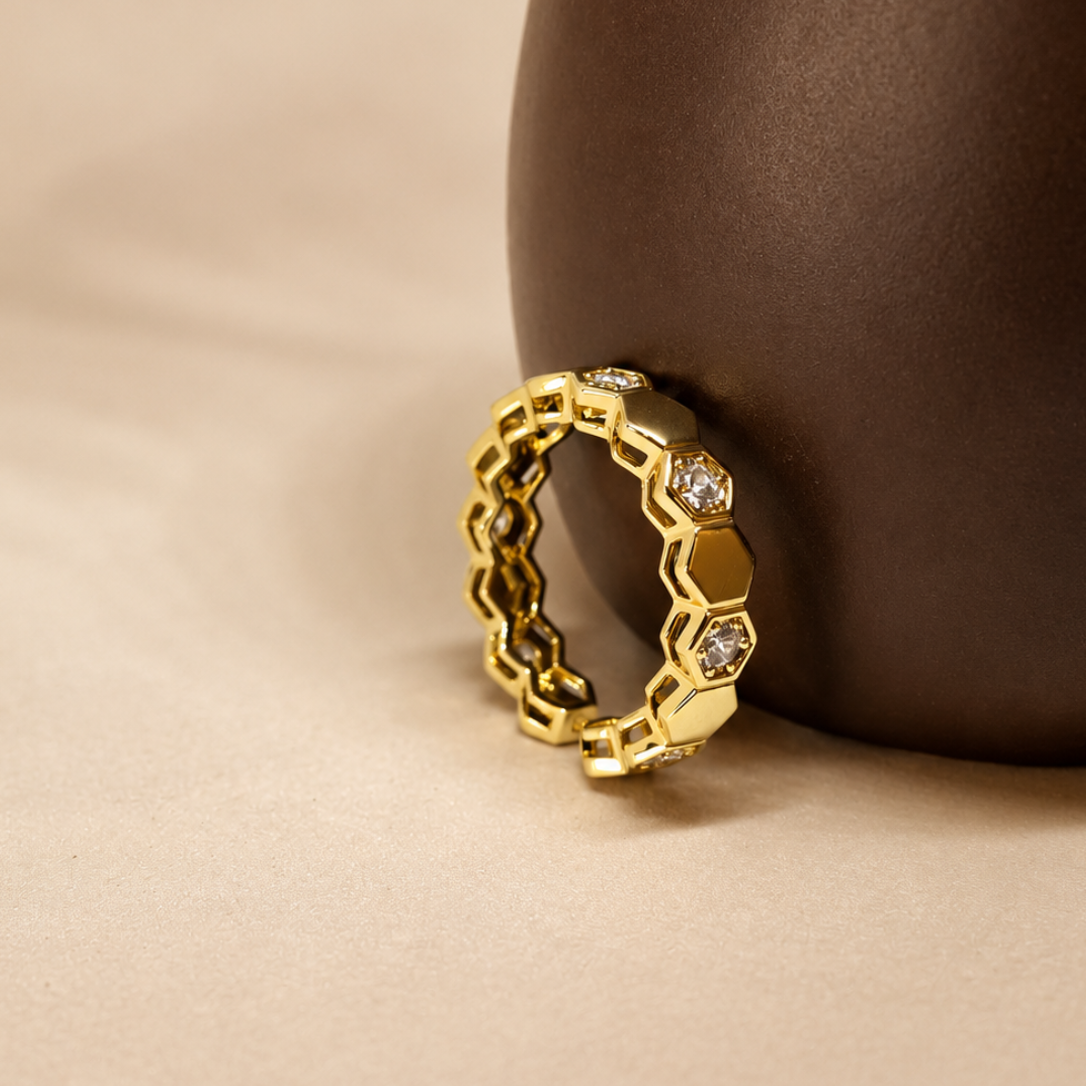 A gold honeycomb-patterned ring with small clear stones, displayed on a dark brown ceramic vase against a beige fabric background.