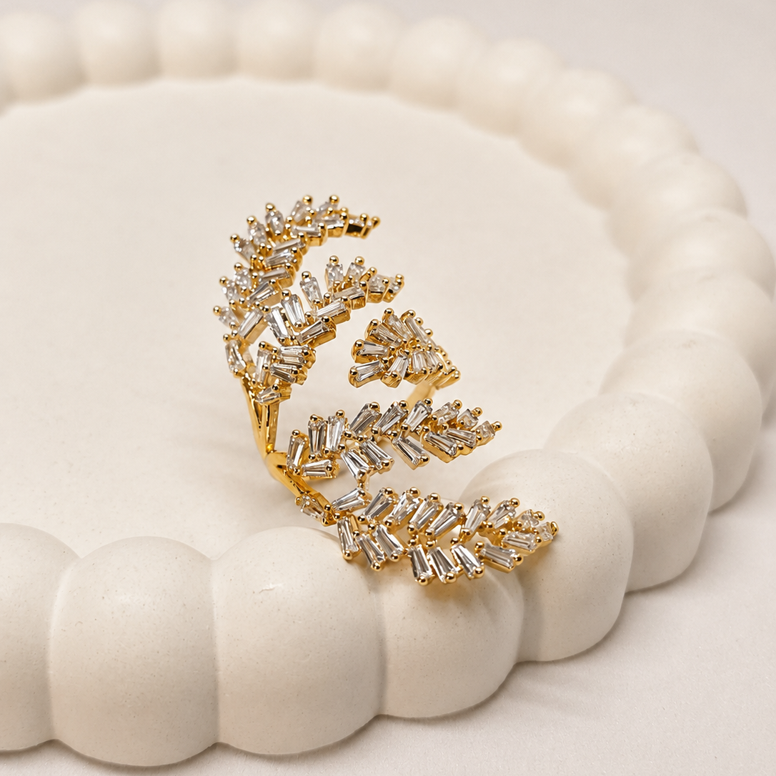 A gold leaf-shaped earring embellished with clear baguette and round crystals, displayed on a white scalloped circular stand against a light beige background.
