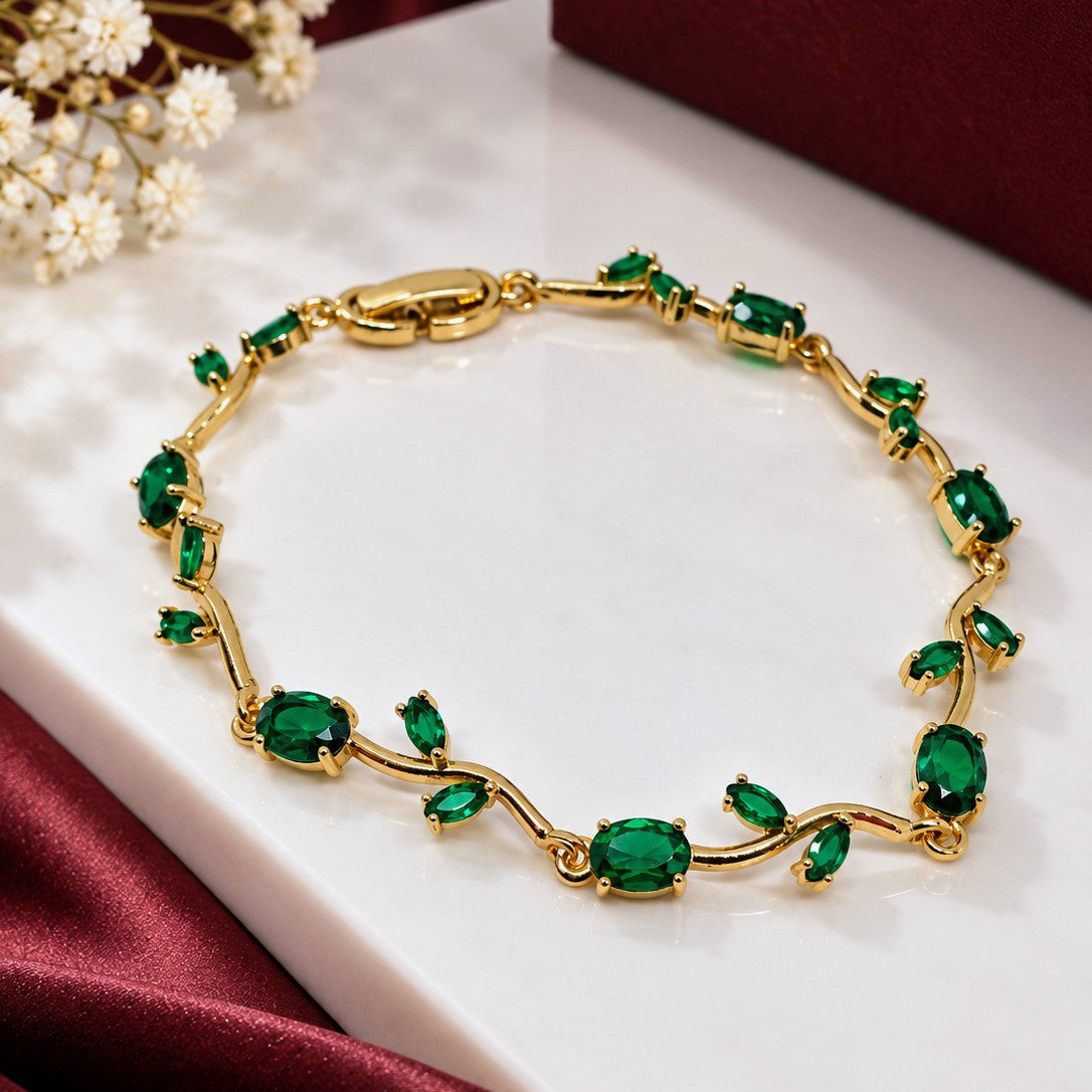 A gold bracelet with multiple emerald-cut green gemstones, displayed on a white marble slab with burgundy satin fabric and small white flowers.