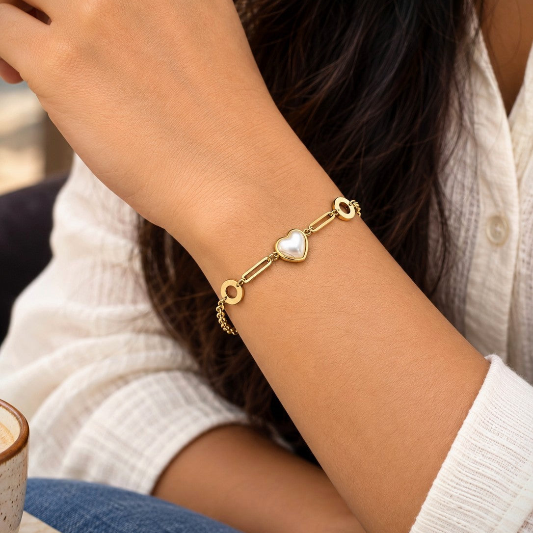 A gold chain bracelet with a central white pearl heart charm, worn on a woman's wrist. The photo is cropped to show the person's hand, arm, hair, and white shirt.