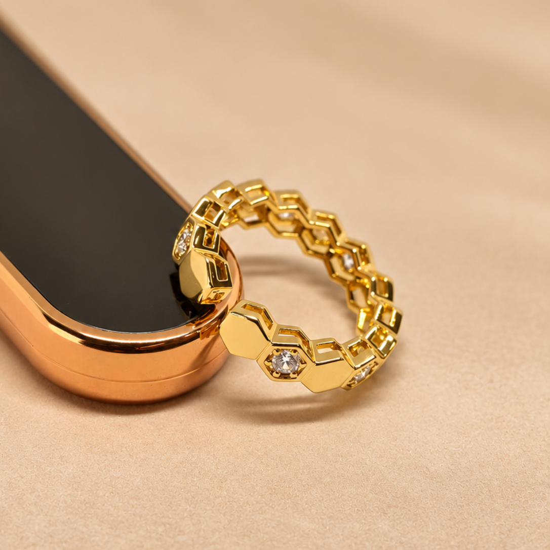 A gold-toned honeycomb-link ring with a small clear gemstone, resting on a rose gold smartphone against a beige fabric background.