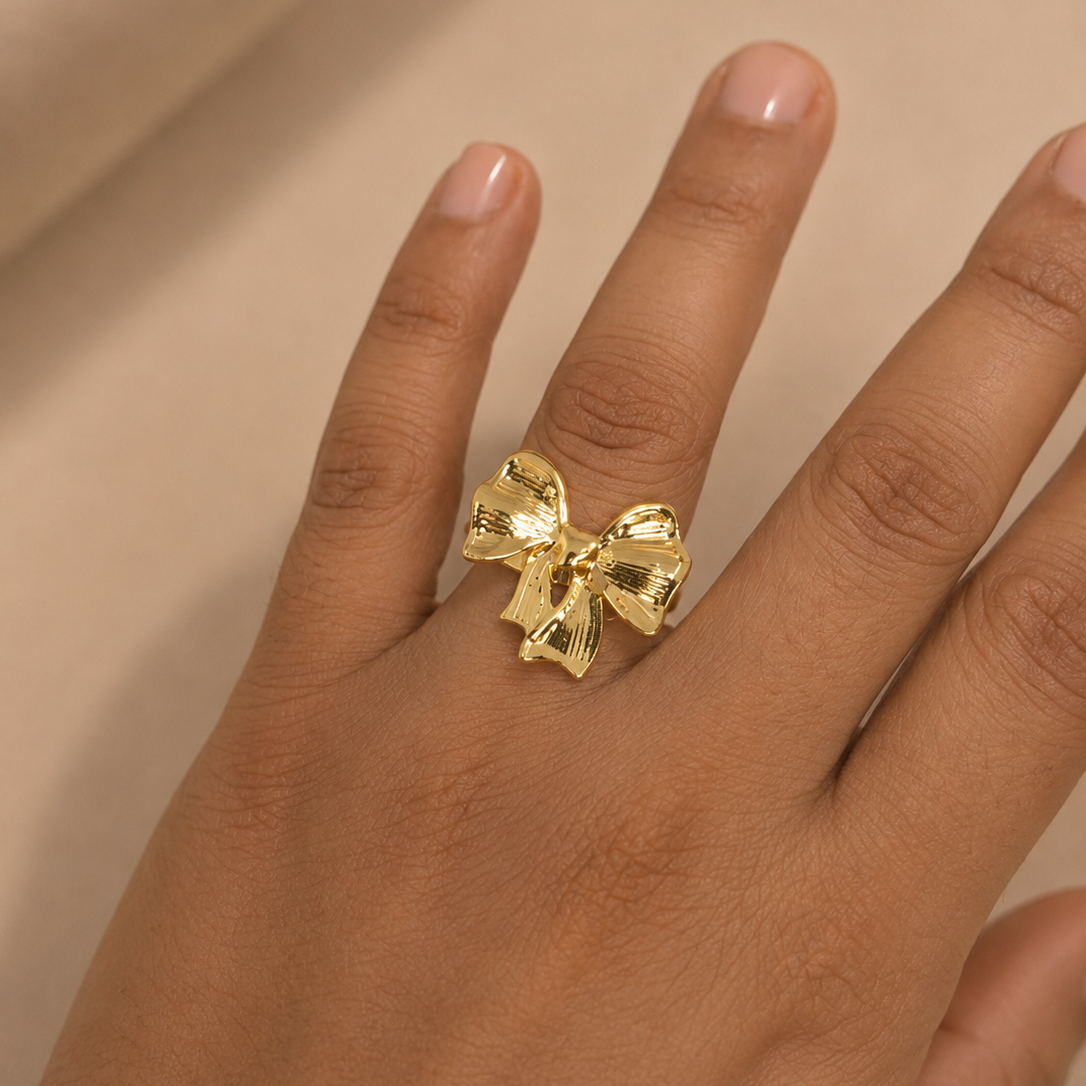 A gold bow-shaped ring worn on a person's finger, shown against a soft beige fabric background.