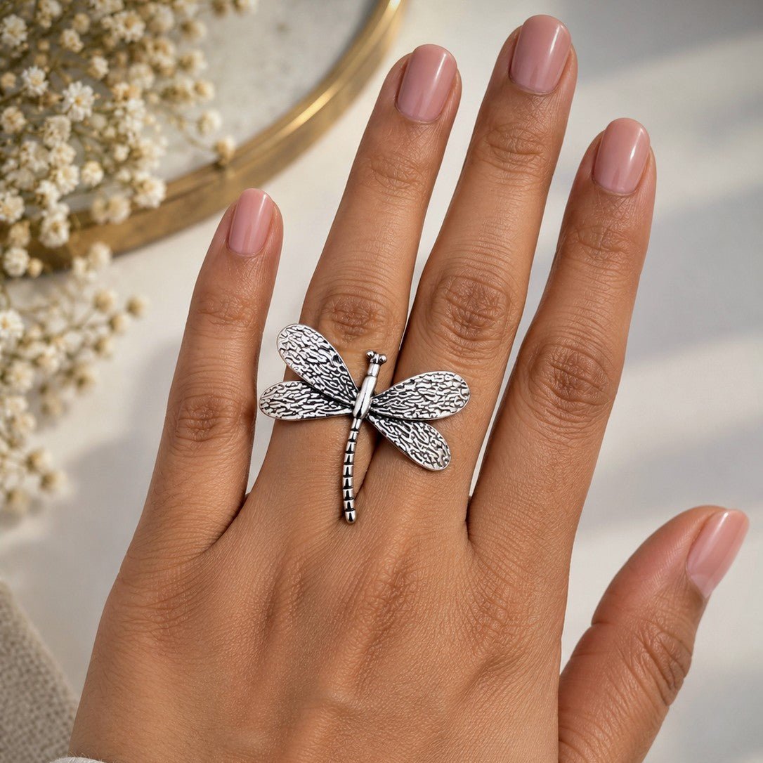 A silver dragonfly ring worn on a hand with pink manicured nails, set against a soft, light background with dried flowers in the corner.