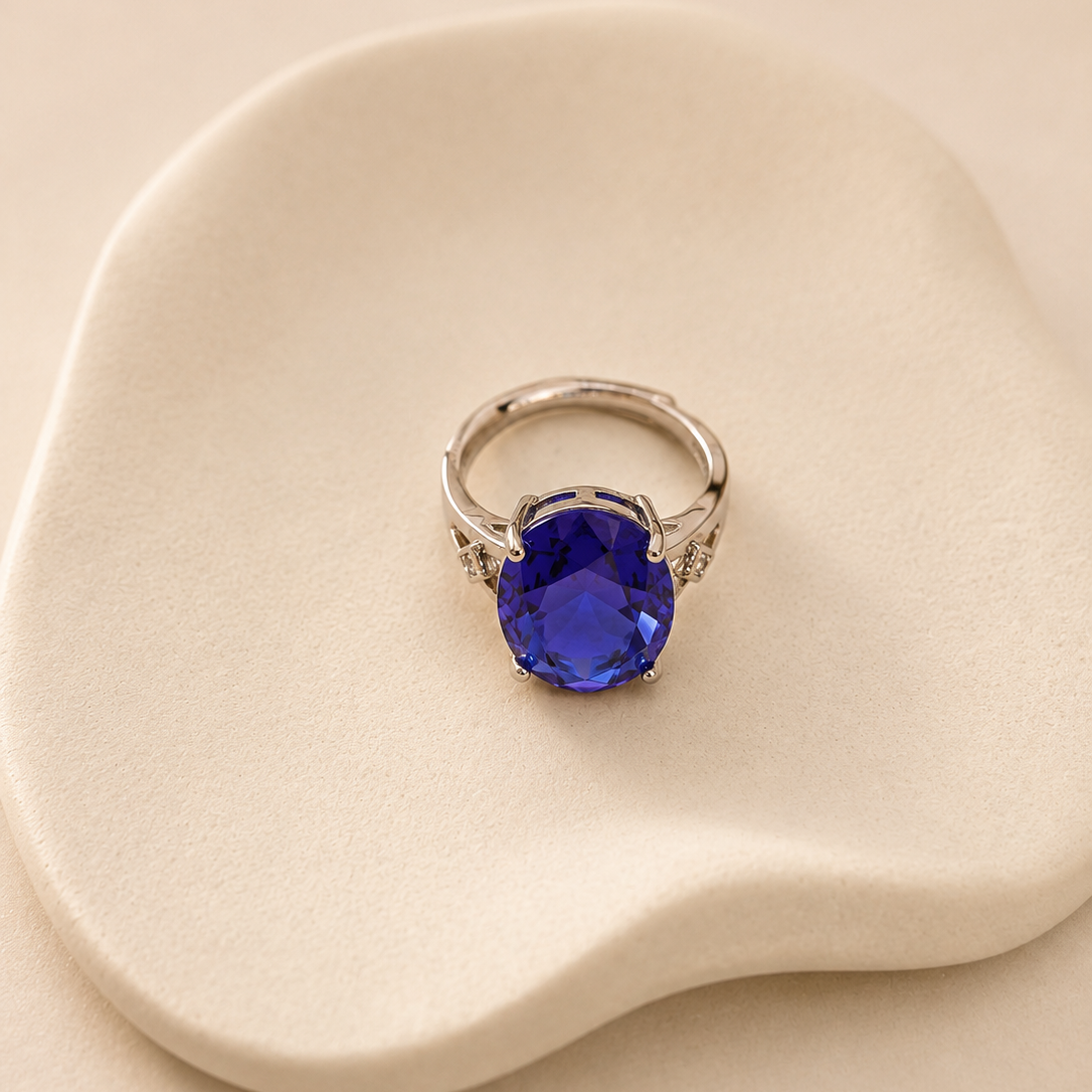 A silver ring with a large oval-cut deep blue gemstone, accented with small side stones, displayed on a beige textured surface.