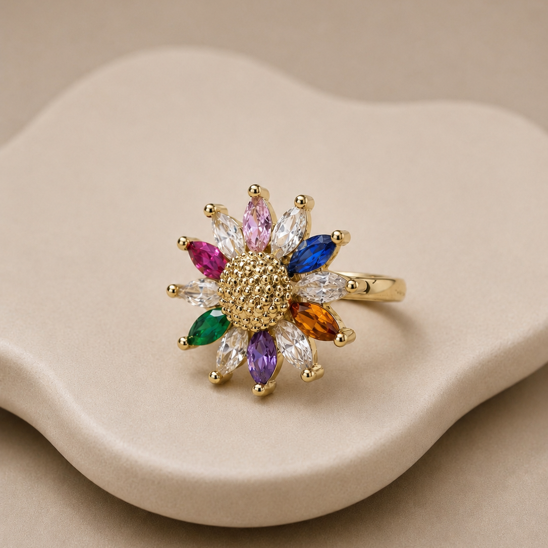 A gold flower-shaped ring with multicolored marquise-cut gemstones, displayed on a beige wavy ceramic stand against a neutral beige background.