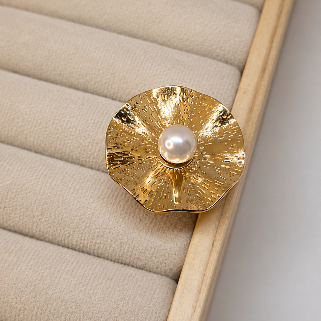 A gold-toned flower-shaped brooch with a textured, hammered finish and a central white pearl, displayed on a beige padded jewelry organizer.