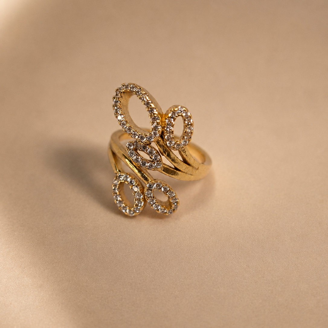 A gold ring with three interlocking oval loops, each encrusted with small clear stones, displayed on a warm beige satin background.