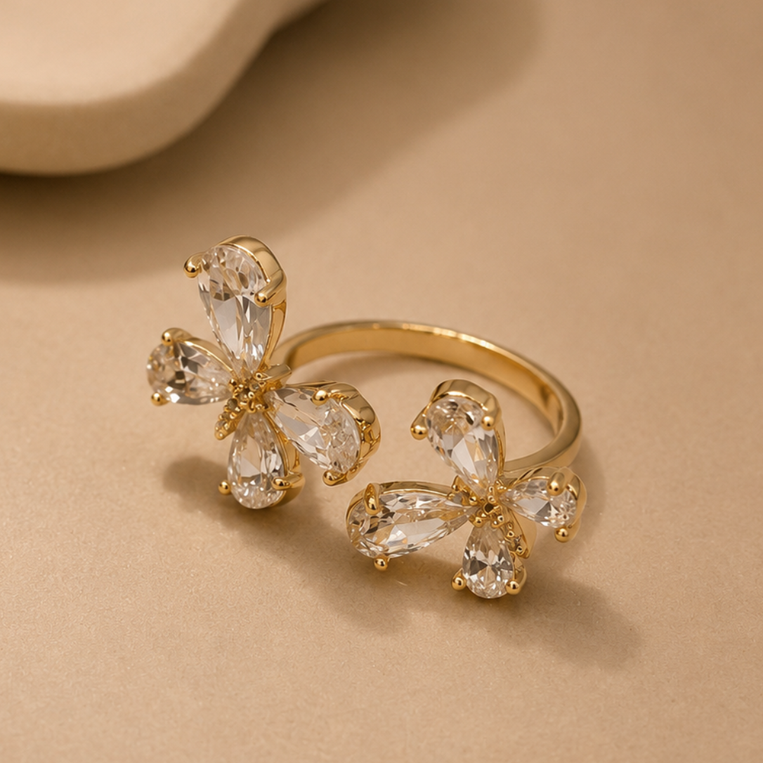 A gold ring featuring two butterfly-shaped clusters of clear teardrop gemstones, displayed on a warm beige surface.
