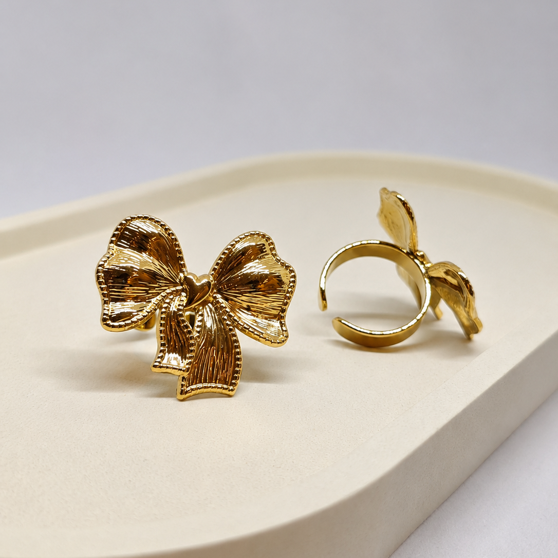 A pair of gold-toned statement earrings shaped like textured bows, one shown from the front and the other from the side/back on a cream-colored display tray against a light gray background.